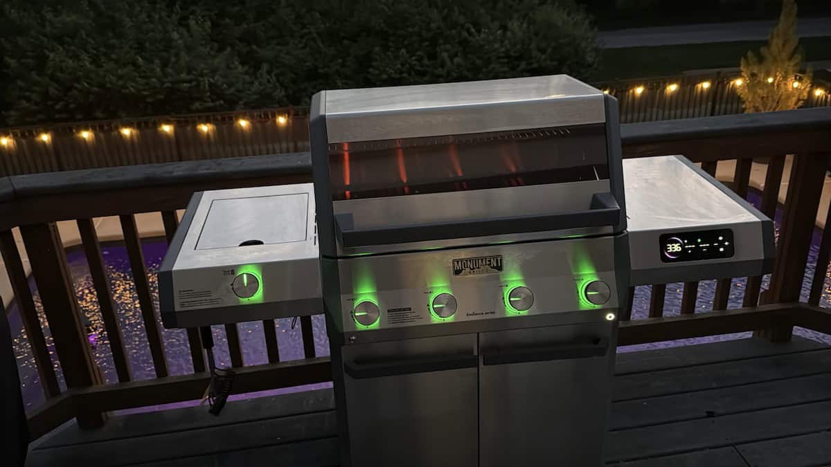Grill on a backyard deck with a lid open, ready for cooking, illuminated by ambient outdoor lighting.