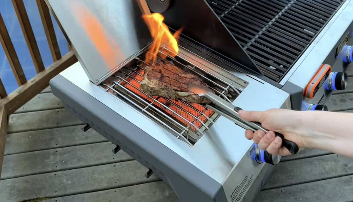 Grilling steak on a gas barbecue with flames and tongs outdoors.